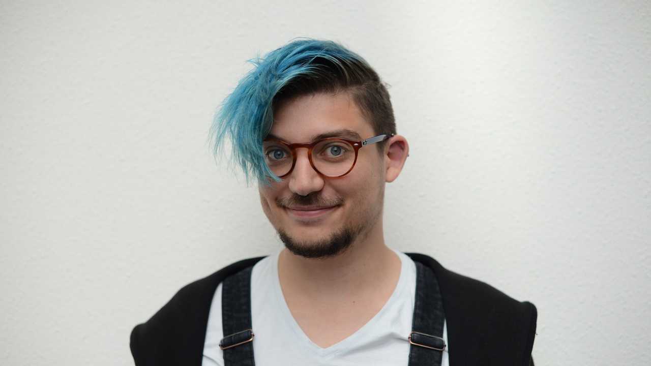 Face portrait of Damien Senger, looking directly at the camera. The person is smiling while wearing an overall outfit. They also have blue hair and several piercings.
